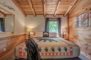 Un dormitorio de cabaña de madera con cama, dos lámparas de noche, una ventana con cortinas y un espejo montado en la pared.