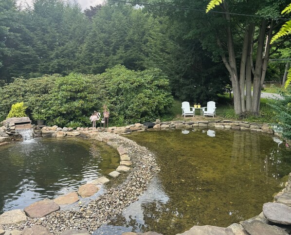A pond with clear water, surrounded by rocks and greenery, with two chairs and a flamingo statue.