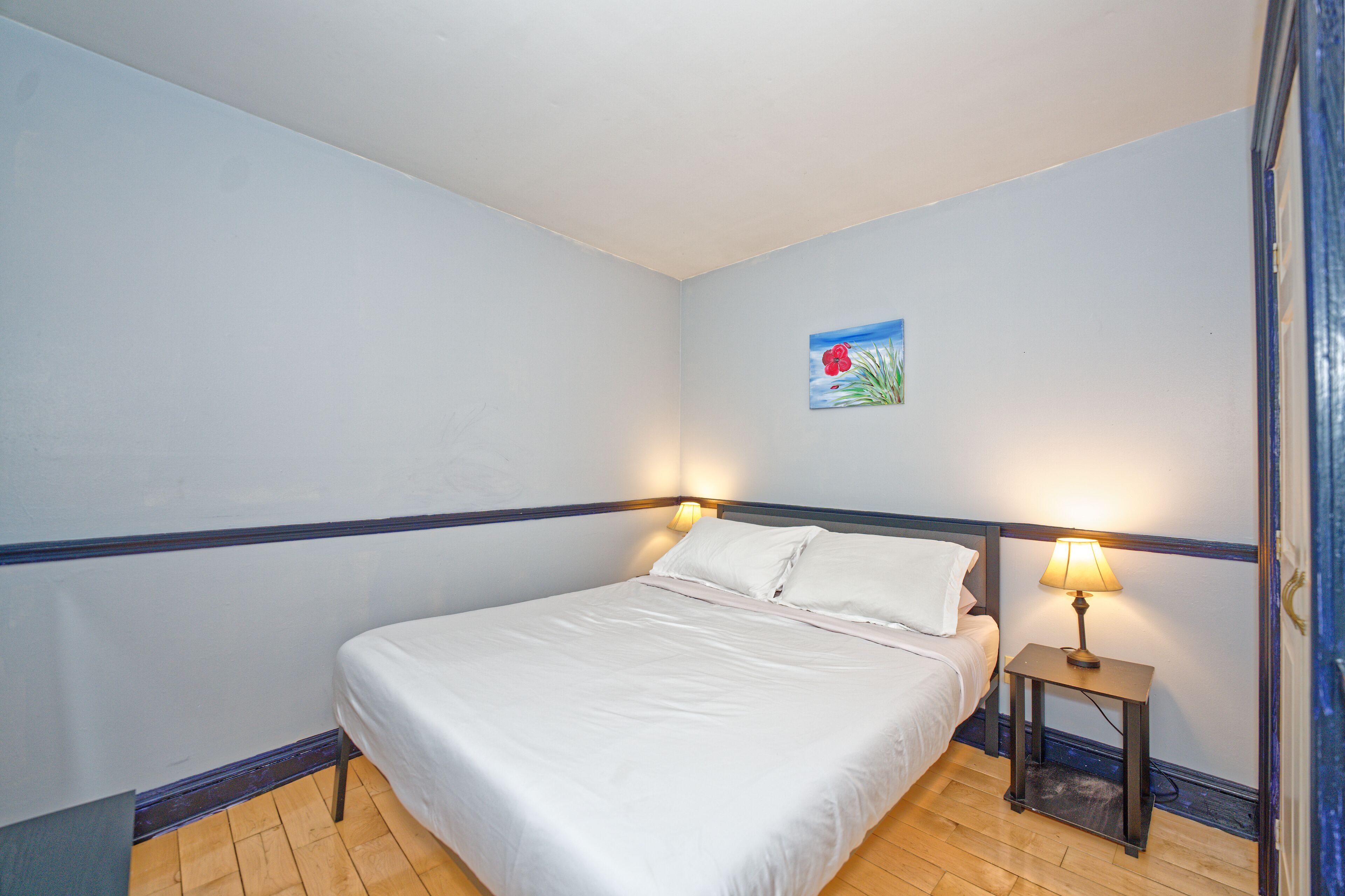 A bedroom with a bed, a nightstand with a lamp, a small painting on the wall, and wooden flooring.