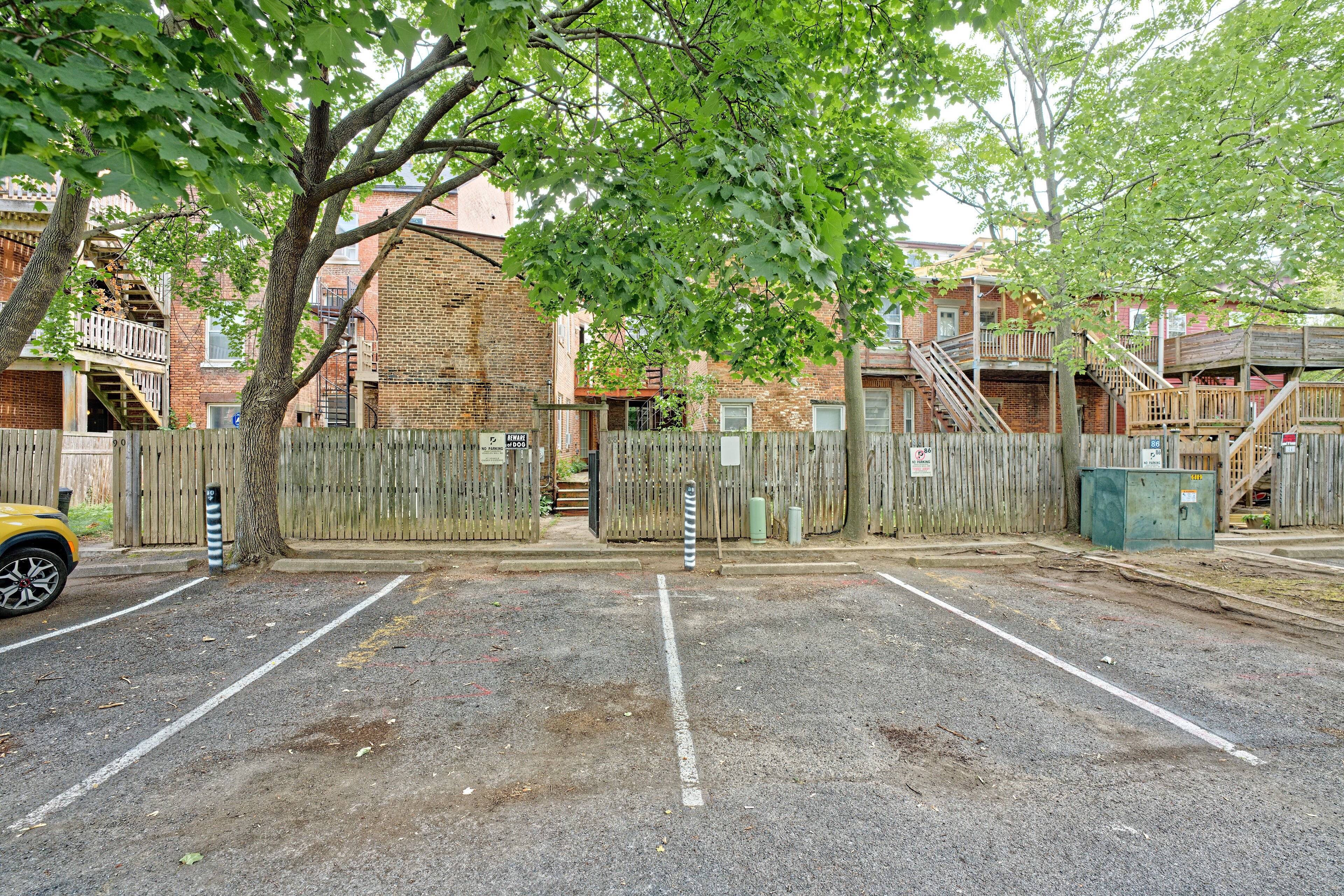 A parking lot with a wooden fence, a yellow car, and a brick building with a fire escape.