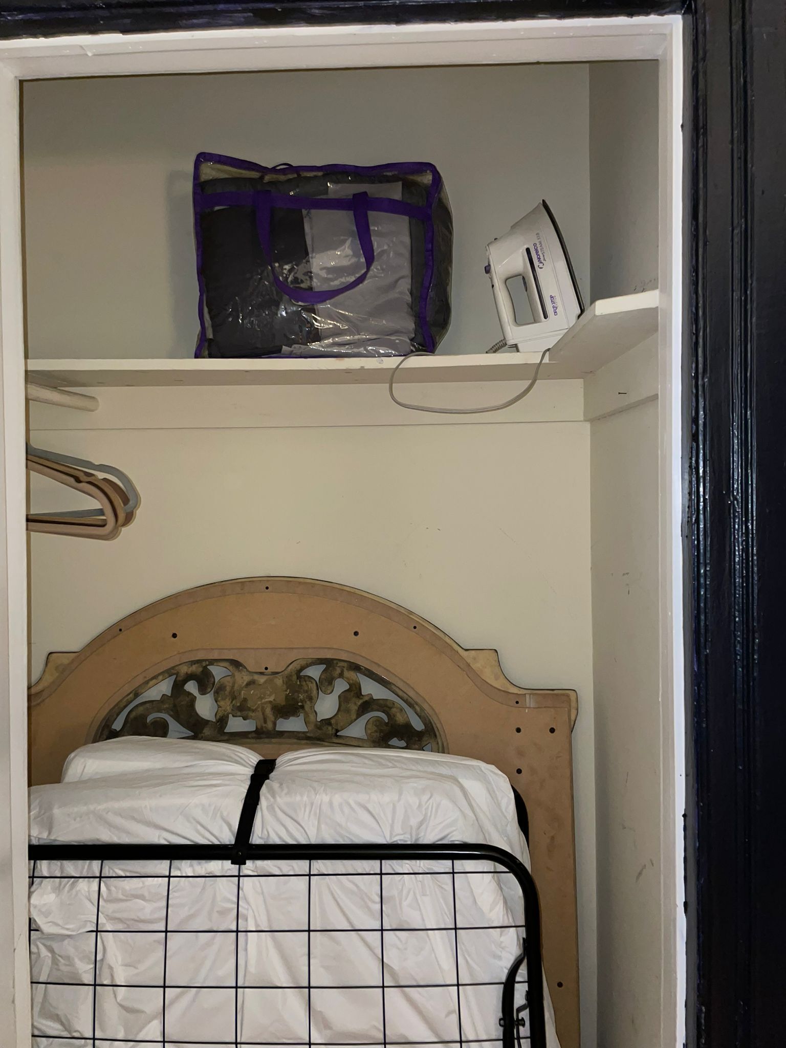 A bedroom with a wooden headboard, a bed with a white mattress and black grid, a purple bag, and a white device on a shelf.