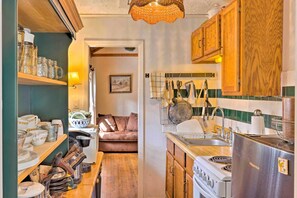 A compact kitchen with wooden cabinets, a stove, and a sink. There are shelves with kitchenware and a dining area visible in the background.