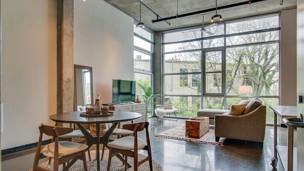 A spacious living area with a round dining table, multiple chairs, a sofa, and a television. Large windows provide a view of the outdoors. - Stylish Gulch Loft Close to Downtown Broadway (Nashville)