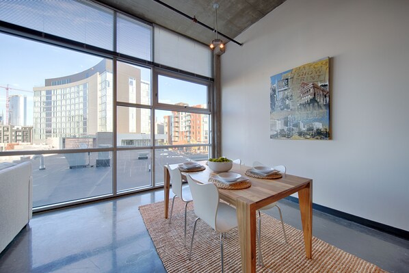 A modern dining area with a large window, a wooden table, white chairs, and a city view.