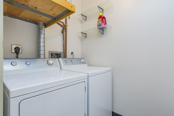 A laundry area with two white front-loading washing machines, a metal exhaust vent, and a shelf with a bottle of detergent.