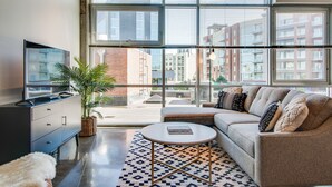 Ein modernes Wohnzimmer mit Sofa, Couchtisch und Fernseher, großen Fenstern mit Blick auf die Stadt und einer Zimmerpflanze.