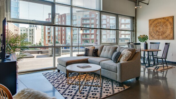 Ein modernes Wohnzimmer mit einem großen Fenster, einem grauen Sofa, einem runden Couchtisch und einem Essbereich mit Stühlen und einem Tisch.