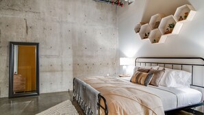Ein modernes Schlafzimmer mit einer Betonwand, einem Bett mit beige-weißer Bettdecke, einem Teppich mit geometrischem Muster und einem einzigartigen sechseckigen Regalsystem darüber.