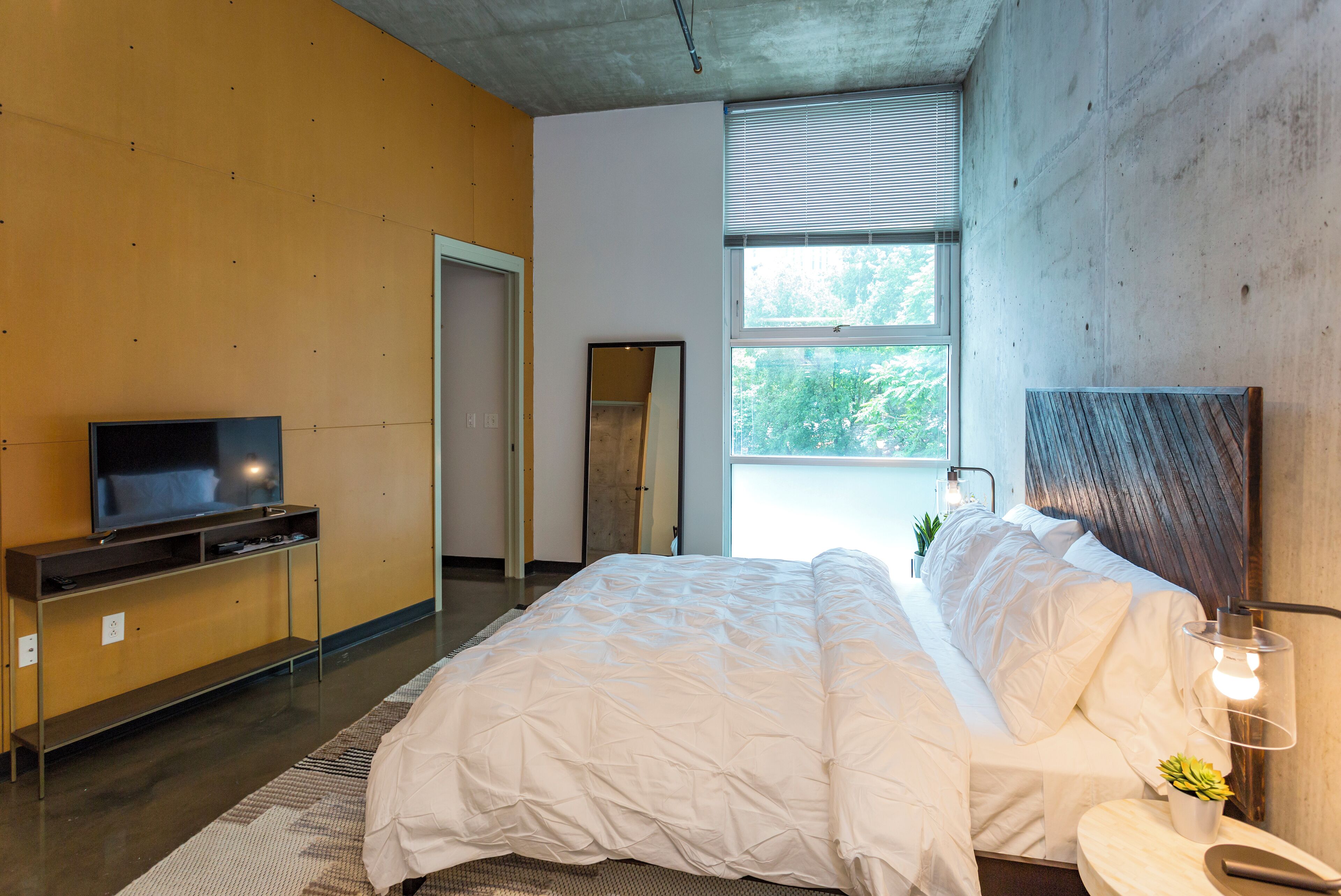Een moderne slaapkamer met een groot bed, een televisie op een houten dressoir en een raam met jaloezieën.