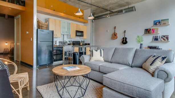 A modern living room with a grey sofa, a wooden coffee table, and a kitchen area with wooden cabinets and a black refrigerator. - Spacious Condo in Downtown Gulch Neighborhood! (Nashville)