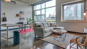 A modern living room with a grey sofa, a coffee table, and a dining area with a metal table and chairs. There is a large window with a view of buildings and a ceiling fan. - Spacious Condo in Downtown Gulch Neighborhood! (Nashville)