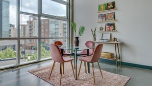 Ett modernt matområde med ett glasbord, rosa stolar och utsikt över byggnader genom stora fönster.
