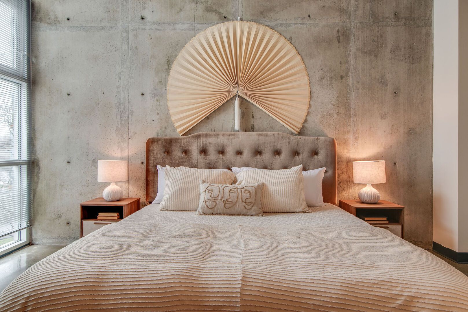 A bedroom with a large bed, two bedside tables with lamps, a round wall decoration, and a concrete wall.