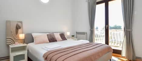 A modern bedroom with a large bed, a bedside table with a lamp, a view of the city through the window, and a cityscape visible outside.