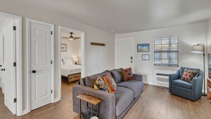 Een moderne woonkamer met een grijze bank, een blauwe fauteuil en een houten salontafel. Achtergrond is een bed en een raam met jaloezieën.