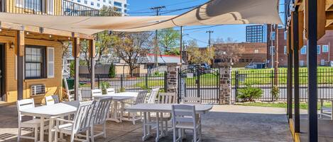Un patio al aire libre con mesas y sillas blancas, un toldo grande y vista a la calle y edificios al fondo.
