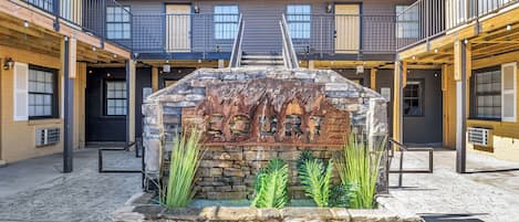 A courtyard with a stone fountain, wooden benches, and a staircase leading to a building with balconies.