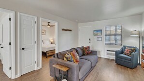 Ein modernes Wohnzimmer mit einem grauen Sofa, einem blauen Sessel und einem Holztisch. Im Hintergrund ist ein Bett zu sehen und ein Fenster mit Jalousien.