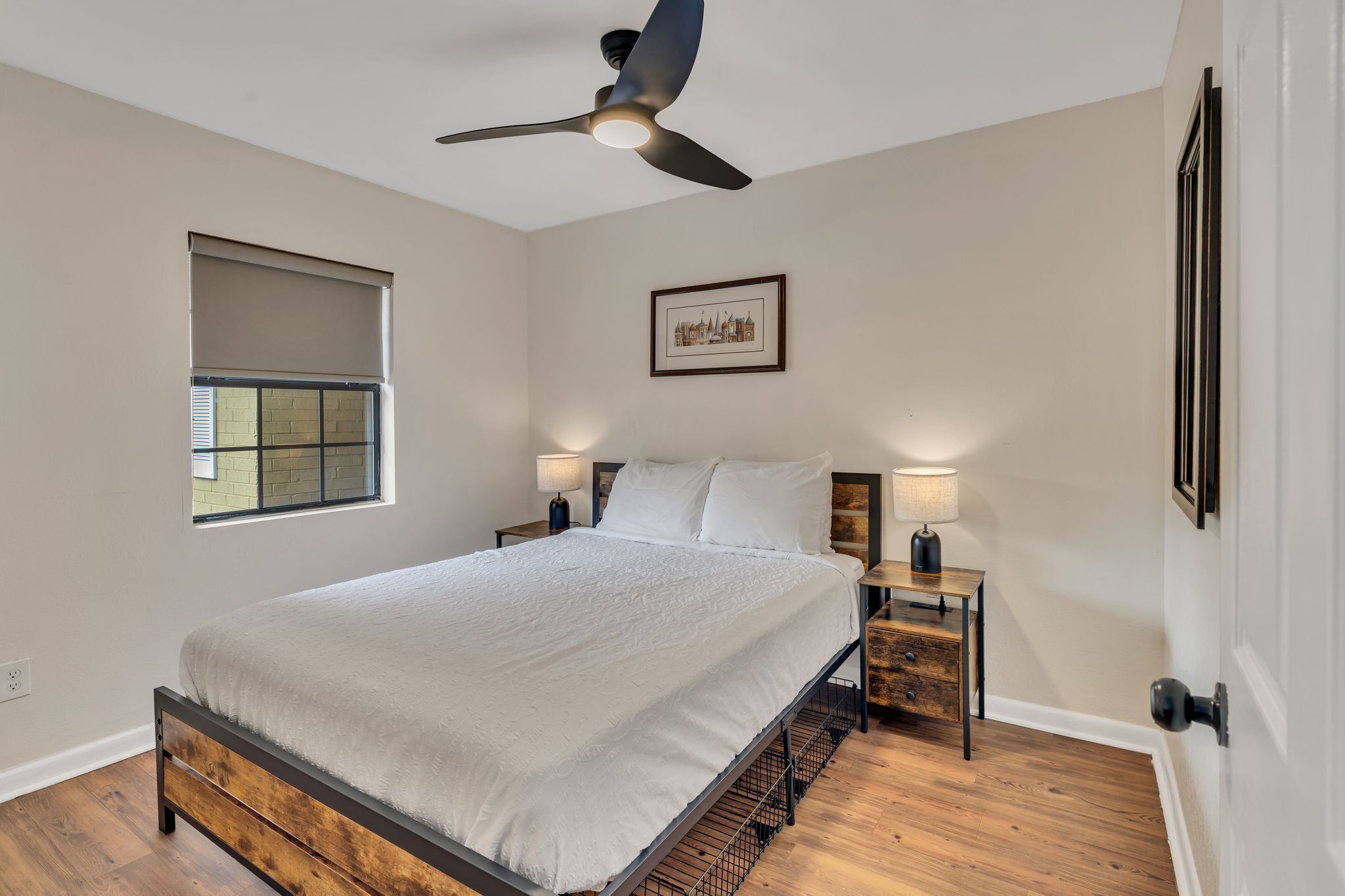 A bedroom with a bed, a ceiling fan, a window with blinds, a nightstand with a lamp, and a framed picture on the wall.