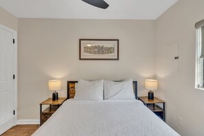Una cama bien hecha con ropa de cama blanca, dos lámparas de noche, un cuadro enmarcado con una vista urbana en la pared y una ventana a la derecha.