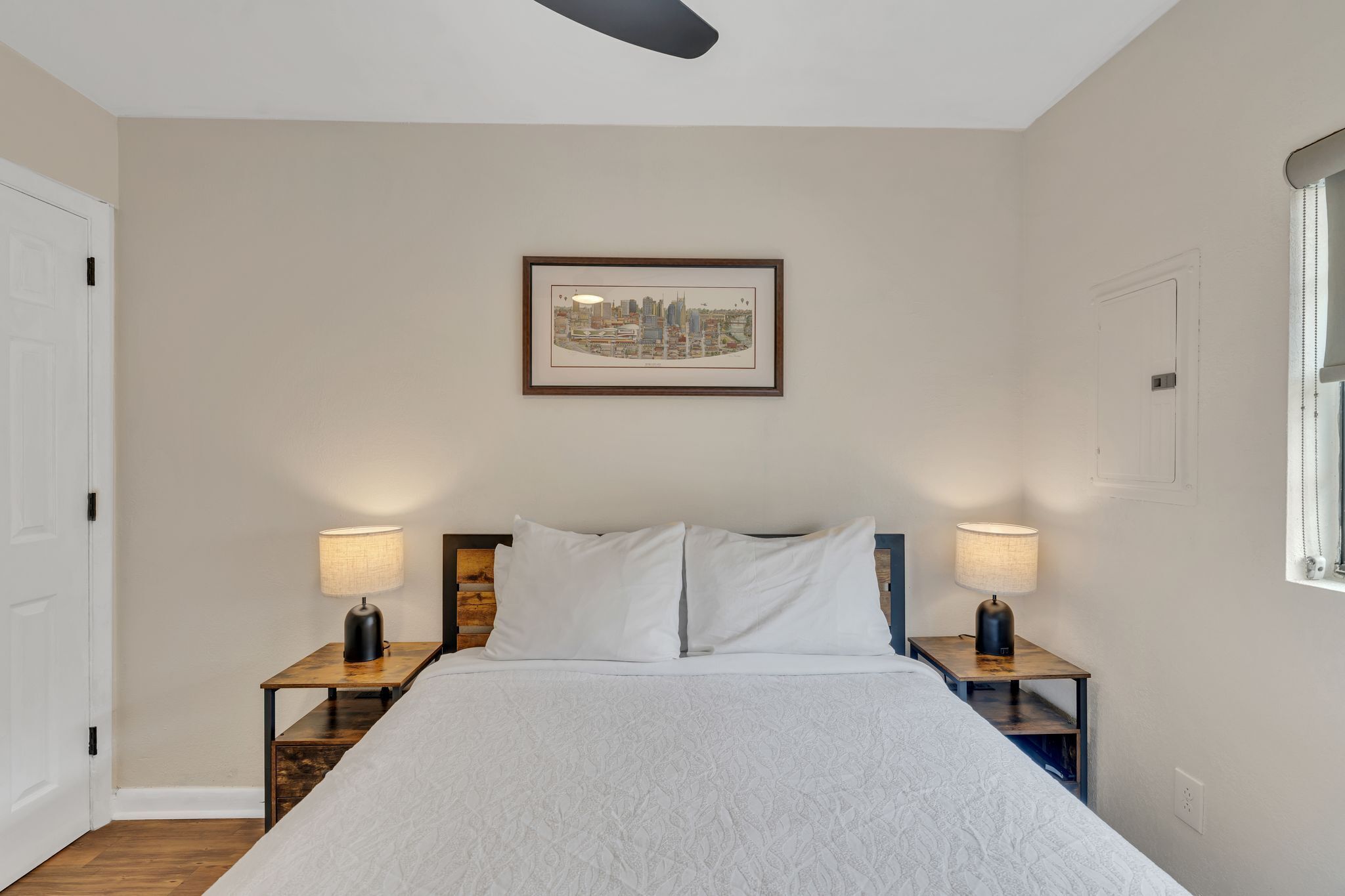 Cama bem arrumada com lençóis brancos, duas luminárias de cabeceira, um quadro com uma vista urbana na parede e uma janela à direita.