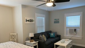 Ein Schlafzimmer mit einem Bett, einem dunkelgrauen Sofa, einem weißen Nachttisch, einem weißen Tisch, einem Deckenventilator und zwei Fenstern mit Jalousien.