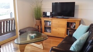 A living room with a glass coffee table, a black leather sofa, a wooden cabinet with books, a television, and a potted plant.