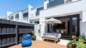 Un patio exterior moderno con terraza de madera, un banco, una mesa con sillas y una planta en maceta.