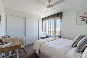 A bedroom with a bed, a desk with books, a chair, a ceiling fan, a window with curtains, and a wall-mounted artwork.