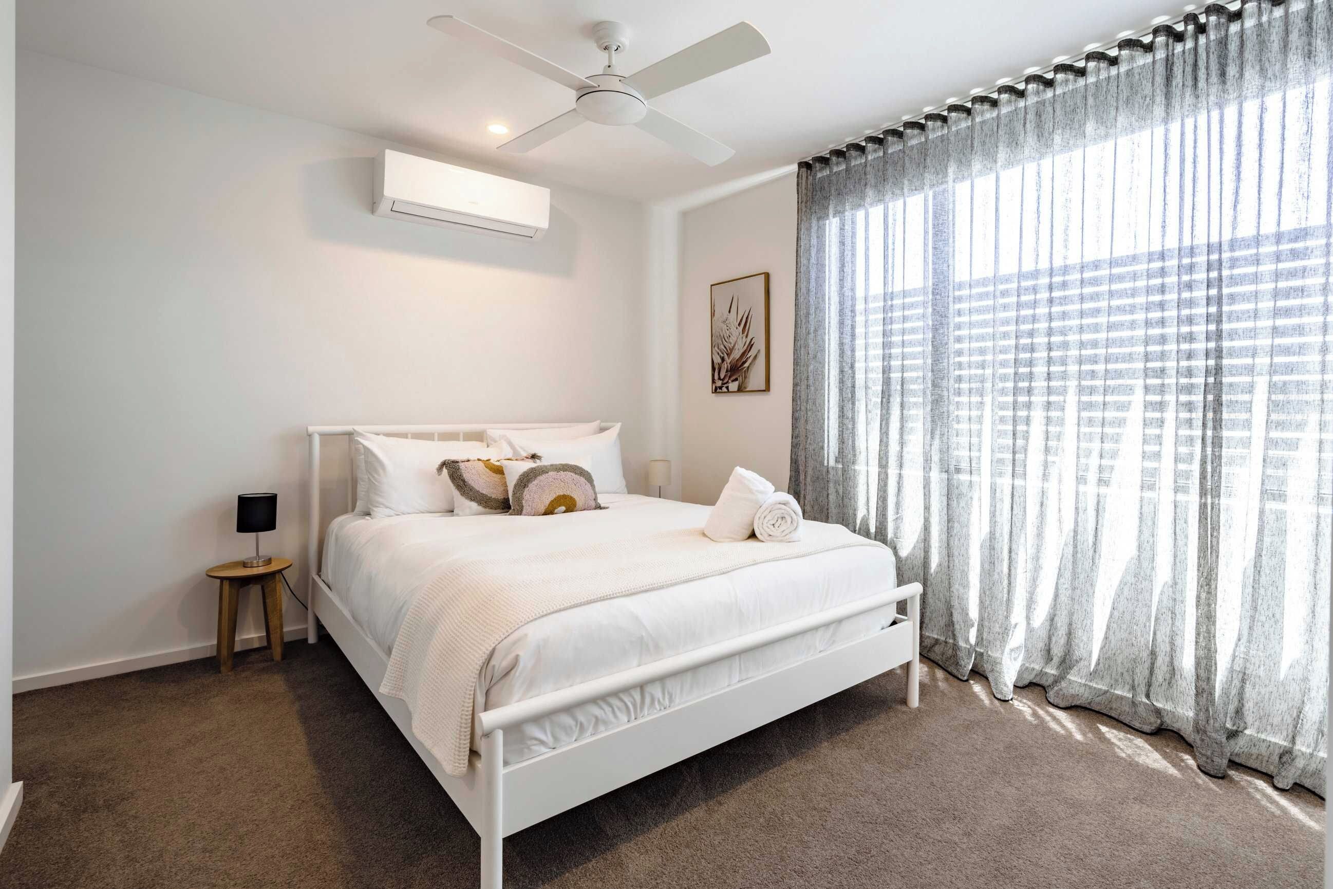 Une chambre à coucher comprenant un lit, un ventilateur de plafond, un climatiseur, une table de chevet avec une lampe et une fenêtre avec des rideaux.