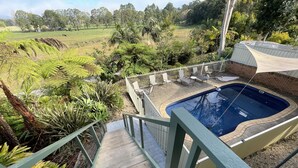 Area piscina con terrazza in legno, circondata da vegetazione lussureggiante e vista su un prato.