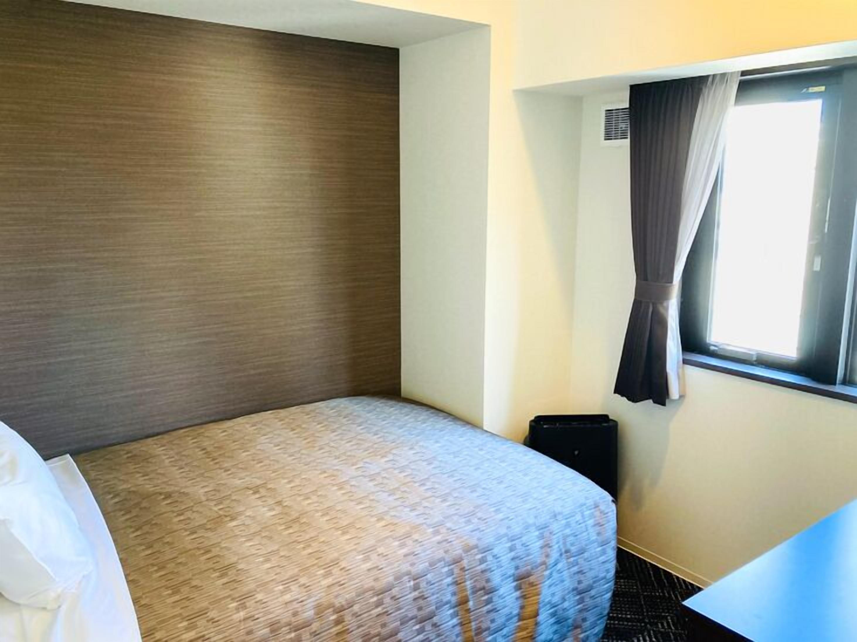 A hotel room with a bed, a window with curtains, a small table, and a wall-mounted air conditioning unit.