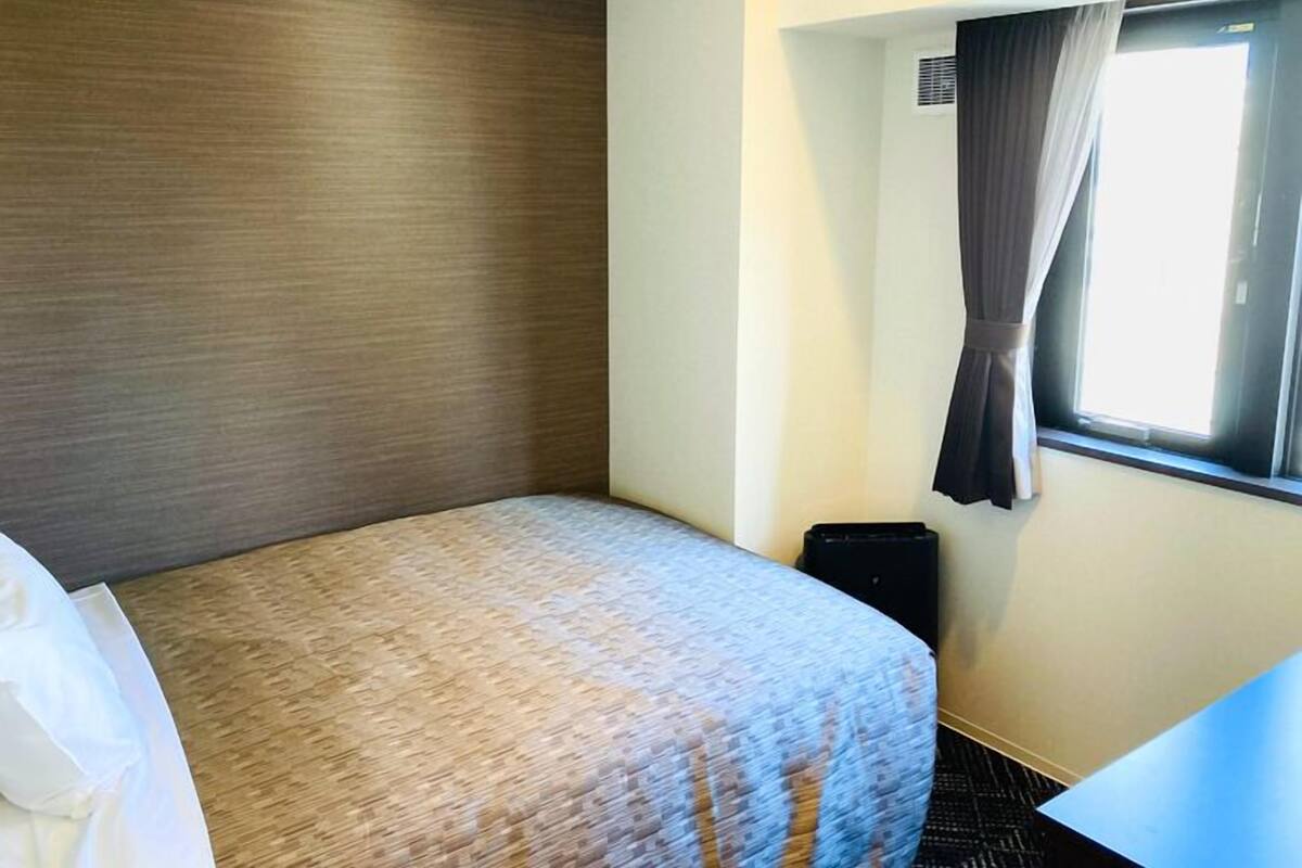 A hotel room with a bed, a window with curtains, a small table, and a wall-mounted air conditioning unit.