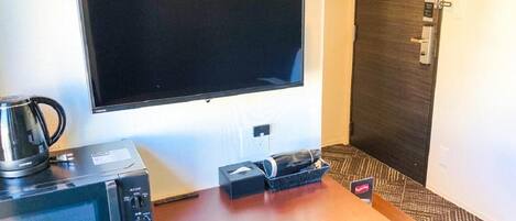 A hotel room with a flat-screen TV mounted on the wall, a coffee maker, a microwave, and a small table with a basket and a remote control.