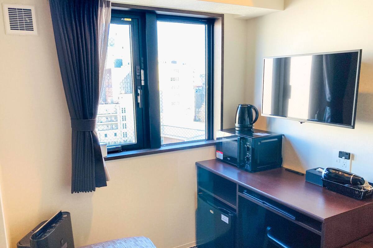 A hotel room with a bed, a desk with a microwave, a coffee maker, a TV, and a window with curtains.