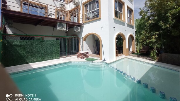 A swimming pool with a clear blue water, surrounded by a white building with arched windows and a green fence. - PALMAS DE CHAHUE C (581 SF) (Crucecita)