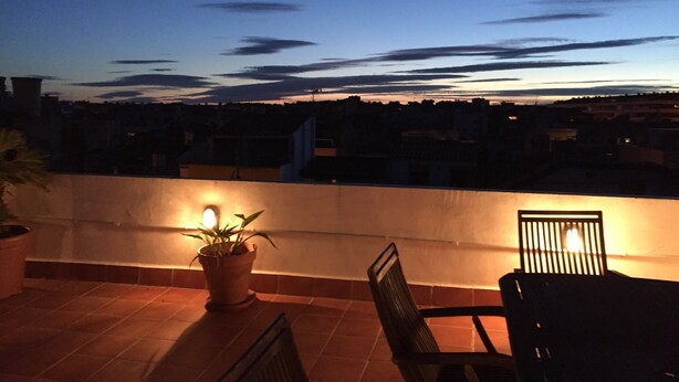A rooftop terrace with a potted plant and two chairs, overlooking a cityscape at dusk.