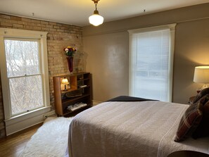 A bedroom with a brick wall, a large window, a bed with a white blanket, a nightstand with a lamp, and a shelf with decorative items. - The Barlow (Hotchkiss)