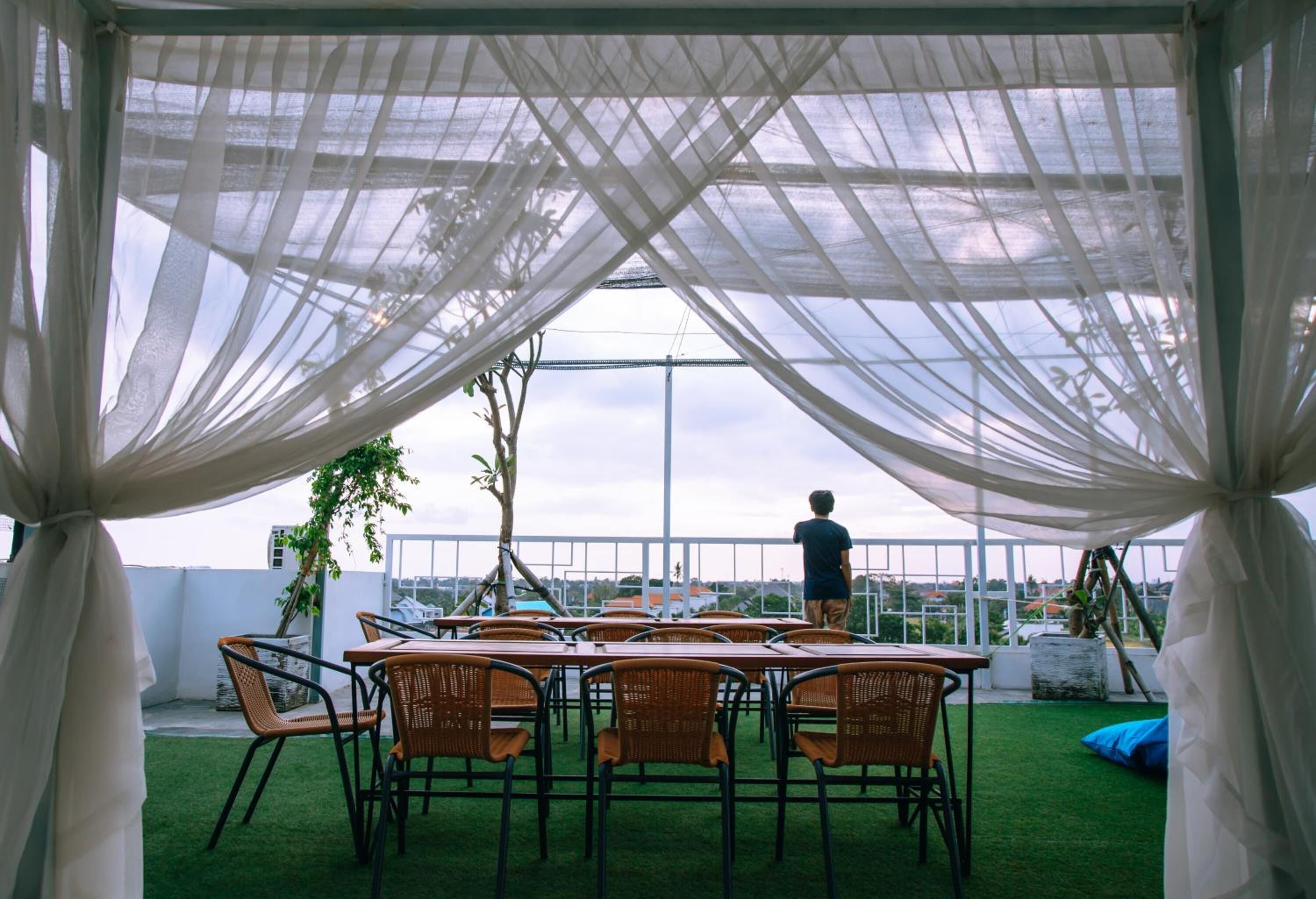 A covered outdoor seating area with tables and chairs, a person standing, and a view of the sky and buildings in the background.
