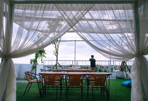 A covered outdoor seating area with tables and chairs, a person standing, and a view of the sky and buildings in the background. - Deva Bali Apartment (Kerobokan)