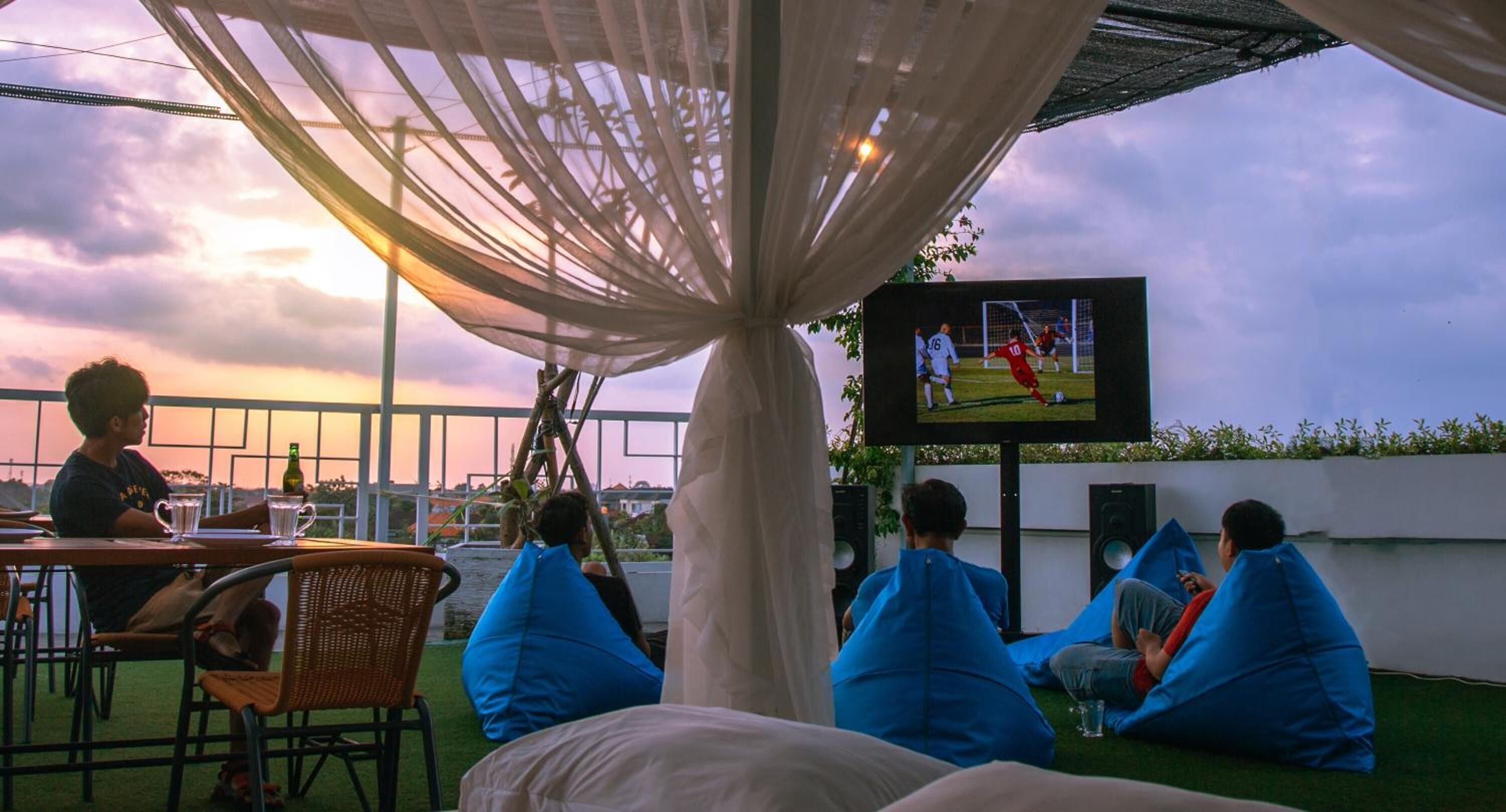 A group of people sitting on bean bags under a canopy, watching a soccer game on a TV screen.