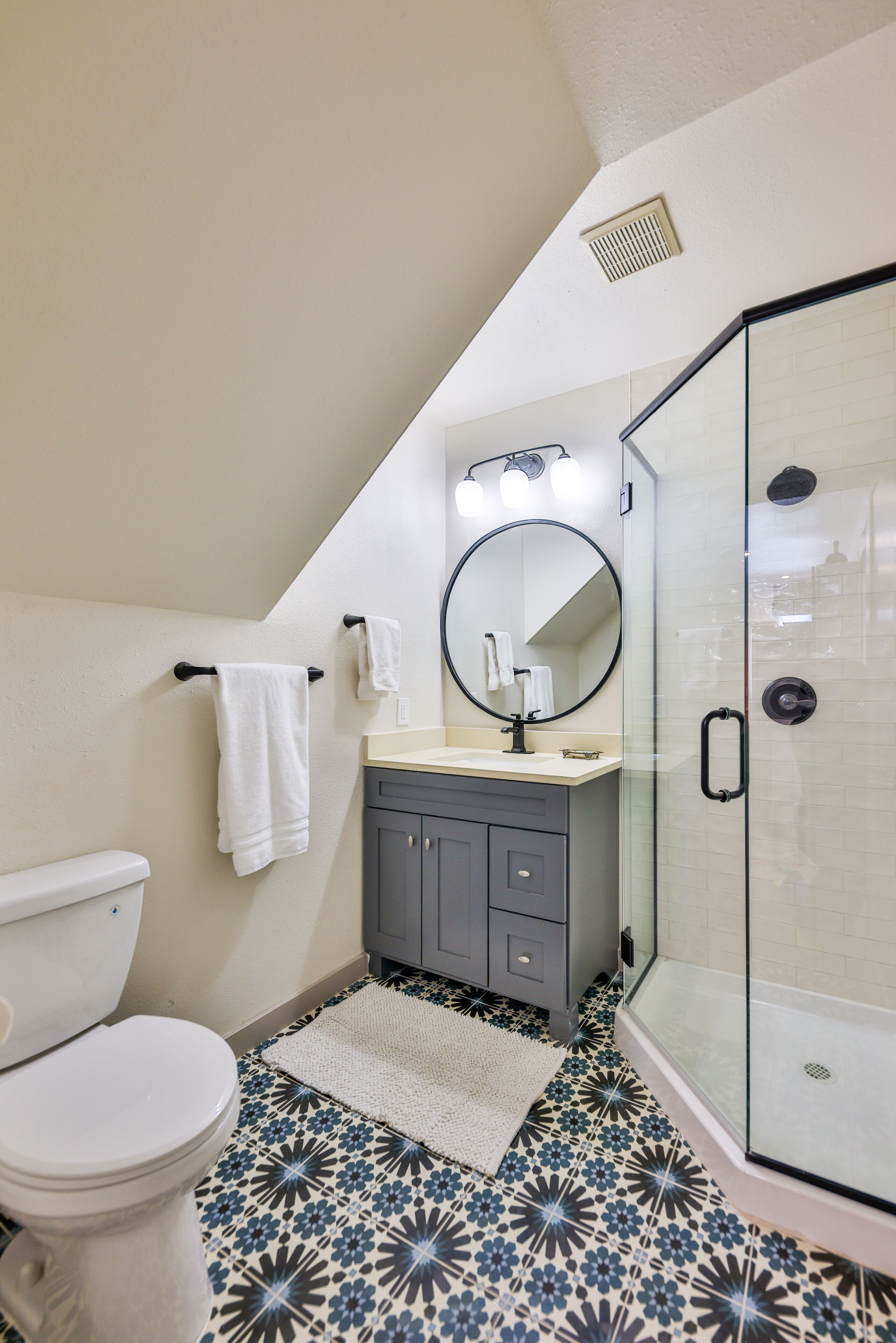 A bathroom with a toilet, a sink with a mirror, a shower with a glass door, and patterned floor tiles.