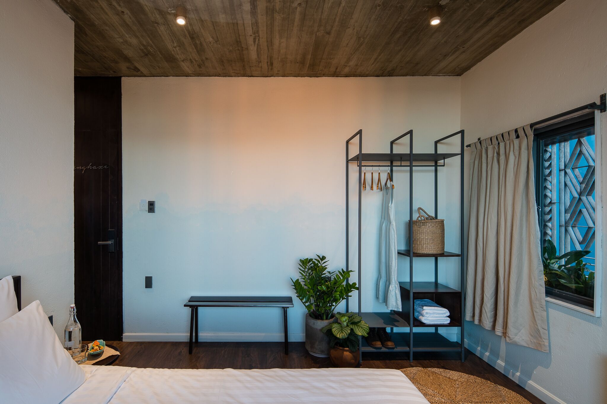 A modern hotel room with a wooden ceiling, a bed with white linens, a bench, a plant, a shelf with towels, and a window with curtains.