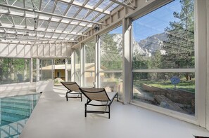 Zona de piscina interior iluminada por el sol, con dos sillones de descanso, grandes ventanales con vistas a la montaña y una estructura de techo de cristal.