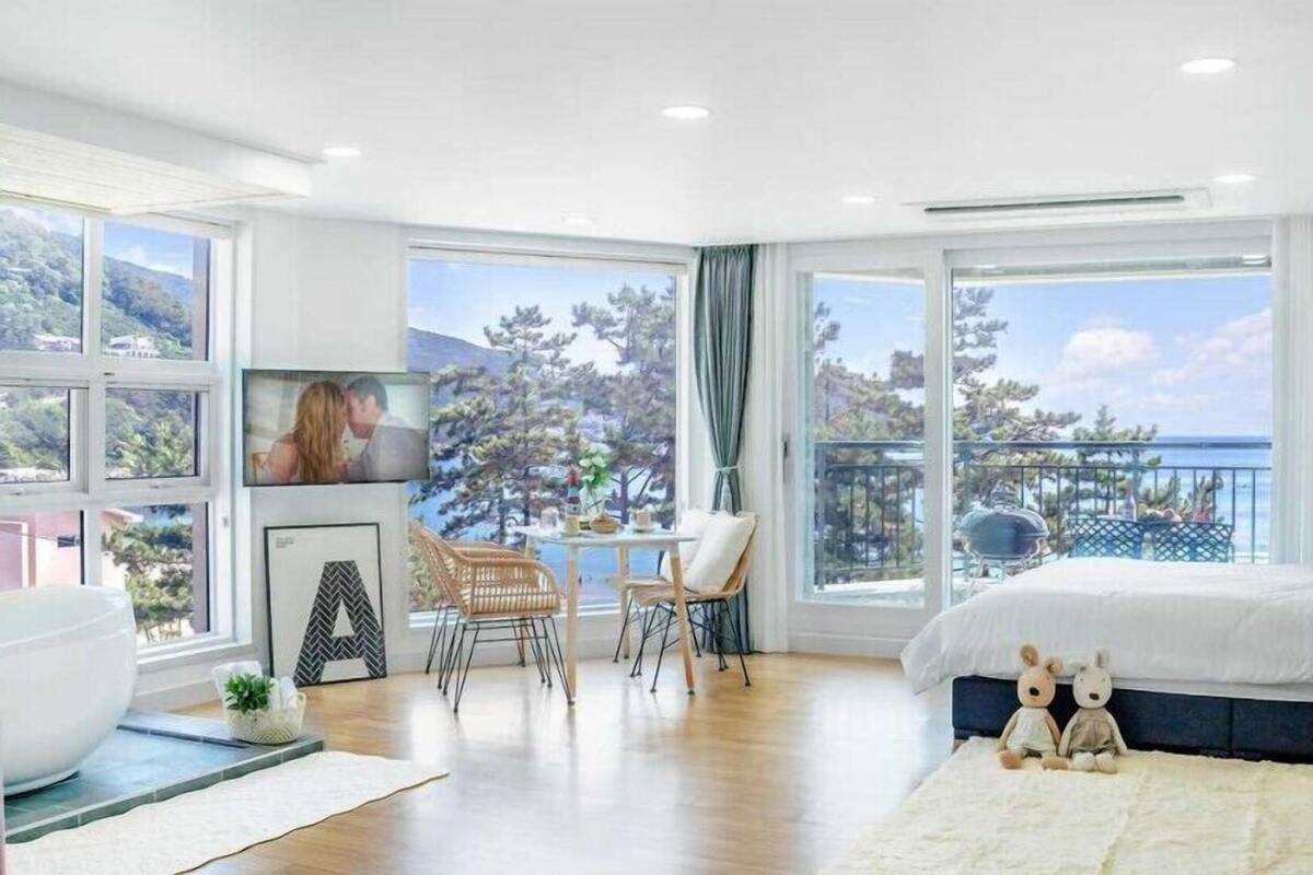 A modern bedroom with a large bed, a seating area with chairs, a dining table, and a view of the sea through large windows.