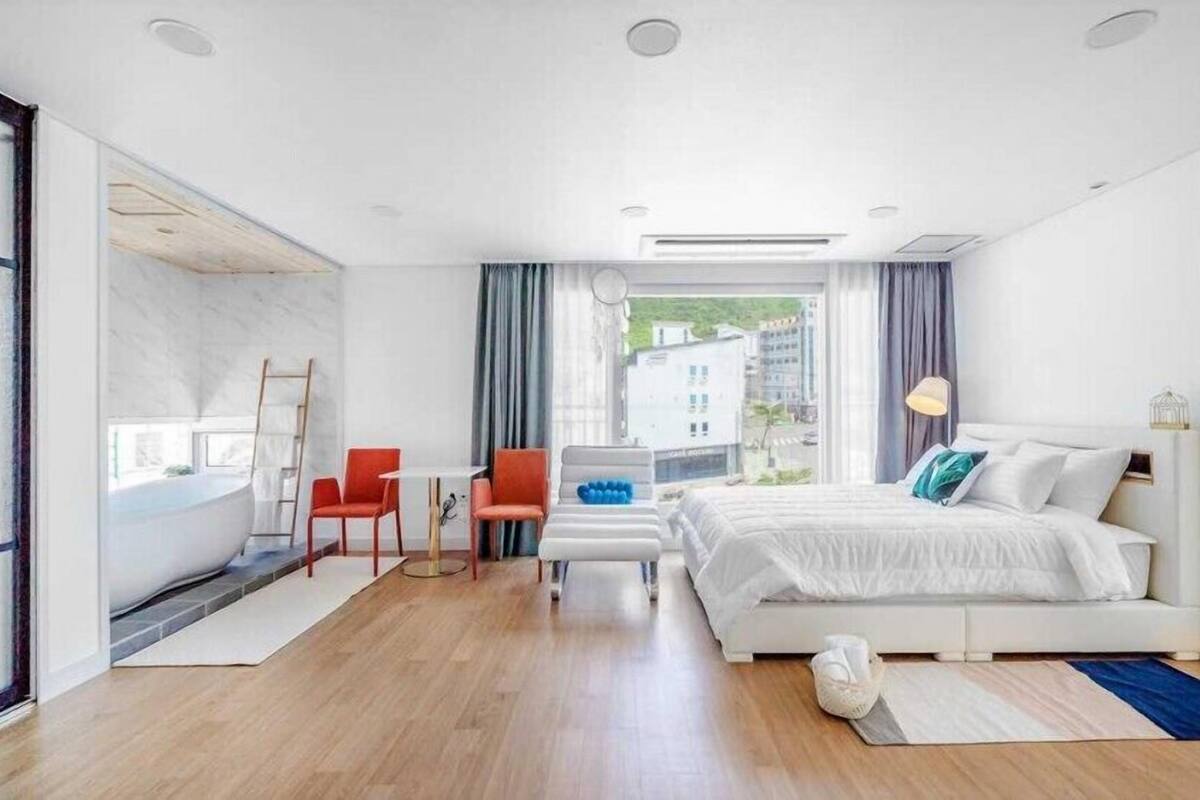 A modern hotel room with a large bed, a desk, and a chair. There is a window with a view of buildings outside.