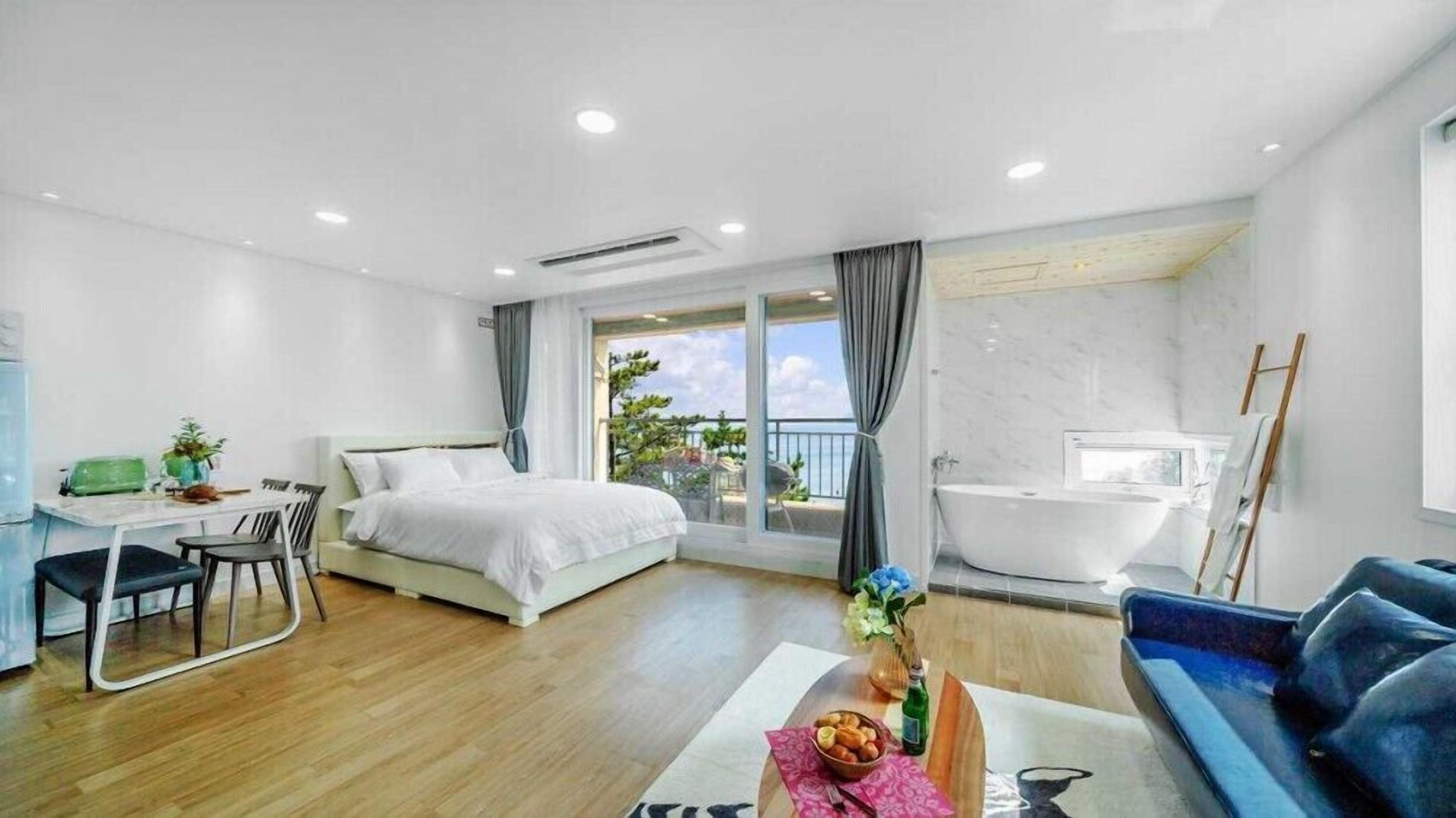 A modern hotel room with a bed, a desk, a bathtub, and a balcony with a view.