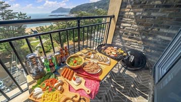 Un balcón con un muro de piedra, una parrilla y diversos alimentos, incluyendo salchichas, verduras y una ensalada.