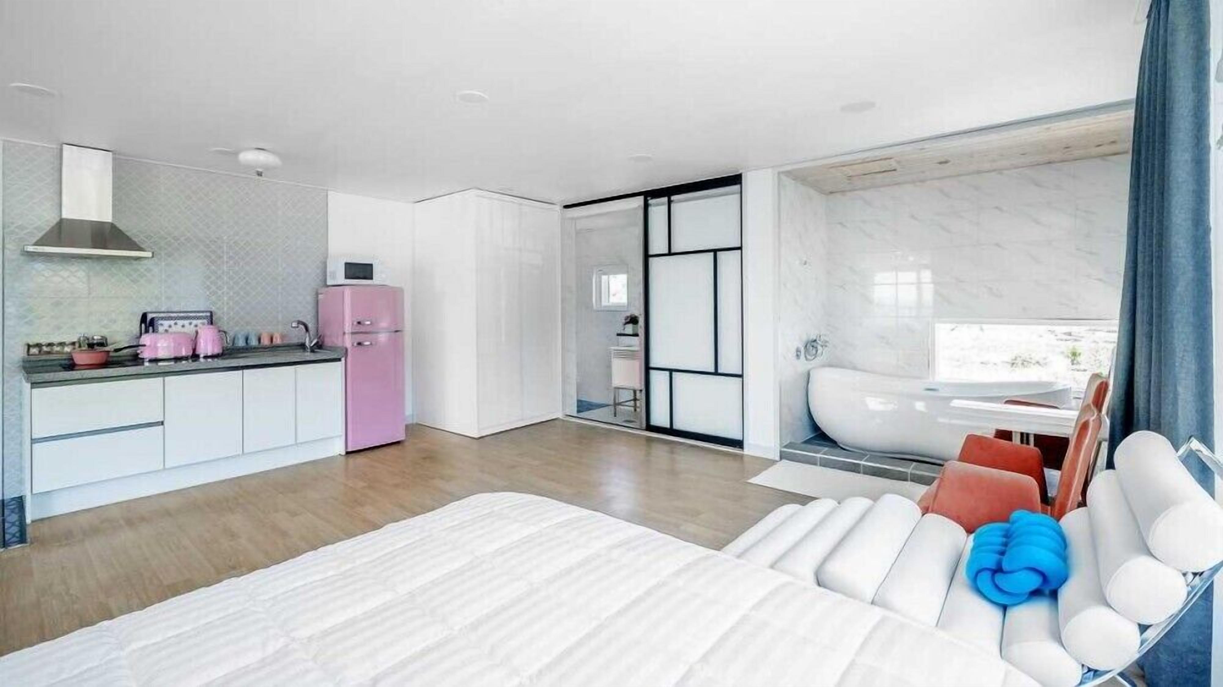 Quarto de hotel moderno com uma cama grande, um espaço de estar com almofadas, uma cozinha com frigorífico rosa e um banheiro com banheira.