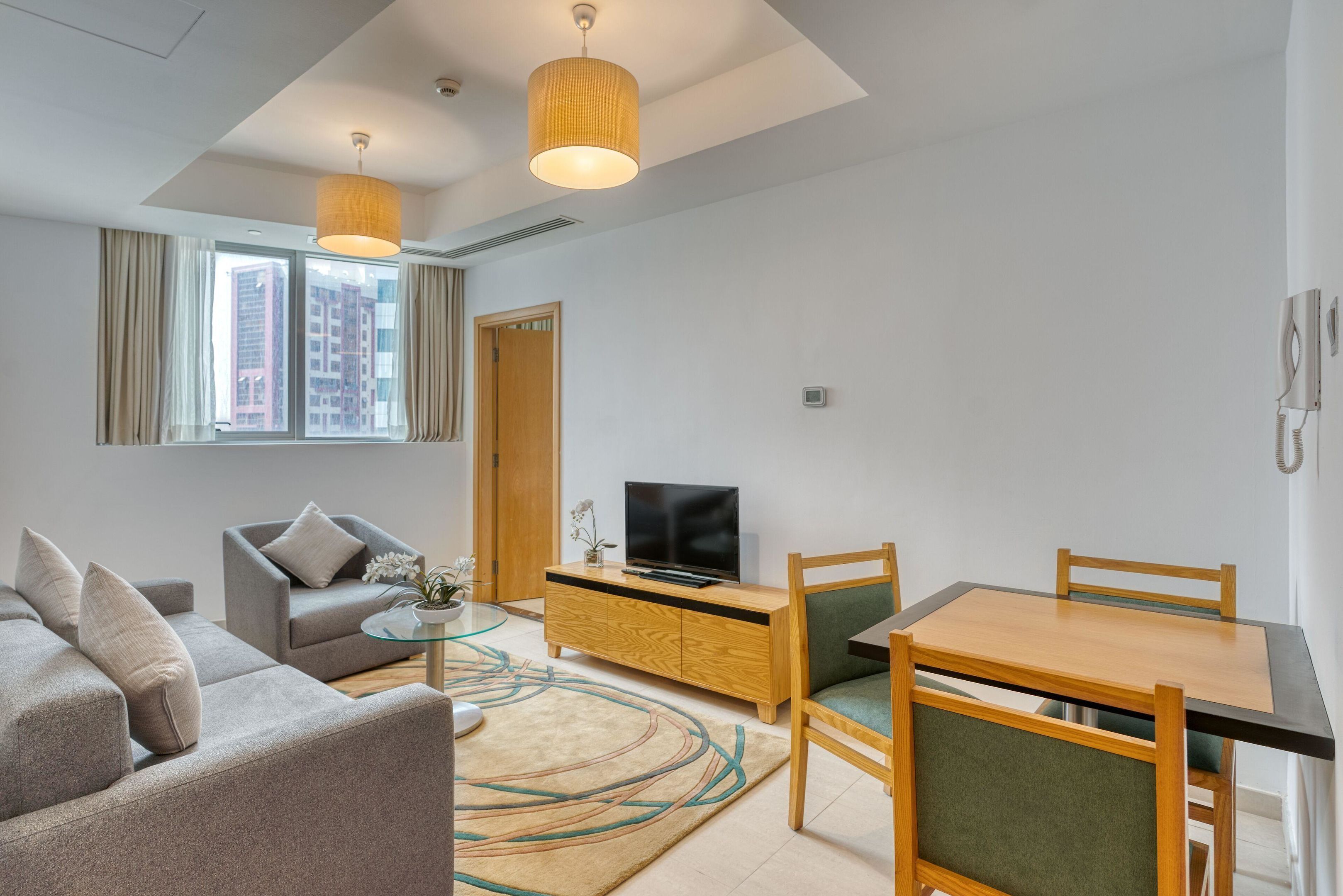 Ein modernes Wohnzimmer mit einer Couch, einem Fernseher auf einer Holzkommode, einem Esstisch mit Stühlen und einem großen Fenster mit Vorhängen.